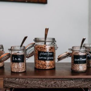 Close-up of labeled glass jars containing various grains and seeds on a wooden table.